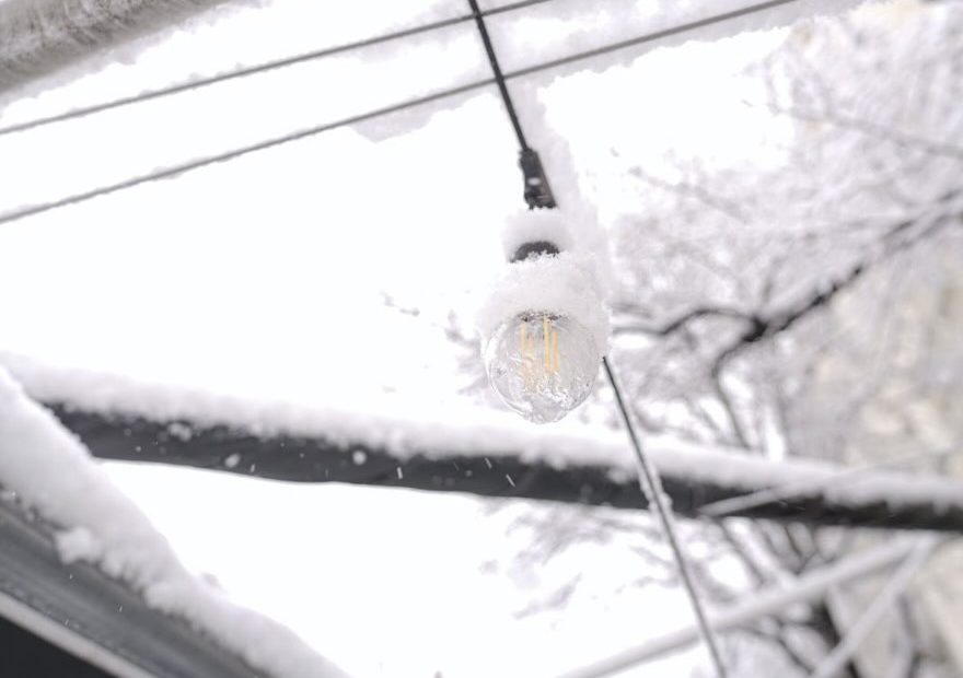 A light bulb hanging from a wire in the snow. The bulb illuminates the wintry scene, creating a contrast between warmth and coldness.